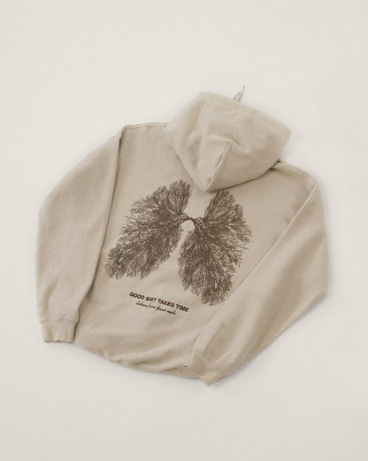 Beiger Kapuzenpullover, flach auf hellem Untergrund liegend. Auf dem Rücken ist eine filigrane dunkelbraune Illustration eines verzweigten Baumes in Form von Lungenflügeln zu sehen. Darunter der Schriftzug „GOOD SHIT TAKES TIME – clothing from planet earth“.
