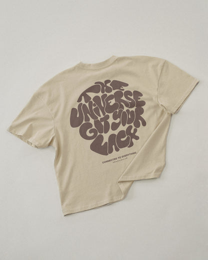 Beiges T-Shirt liegt flach mit dem Rücken nach oben. Der großflächige Druck zeigt den Schriftzug „THE UNIVERSE GOT YOUR BACK“ in verspielten, runden Buchstaben. Darunter steht in kleiner Schrift „CONNECTED TO EVERYTHING – clothing from planet earth“.