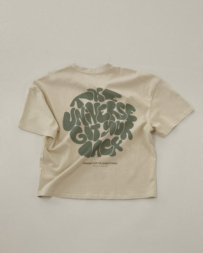 Beiges T-Shirt liegt flach auf hellem Untergrund. Auf der Rückseite ist in großflächiger, dunkelgrüner Schrift der Slogan „THE UNIVERSE GOT YOUR BACK“ im verspielten, organischen Stil gedruckt. Darunter steht „CONNECTED TO EVERYTHING – clothing from planet earth“.