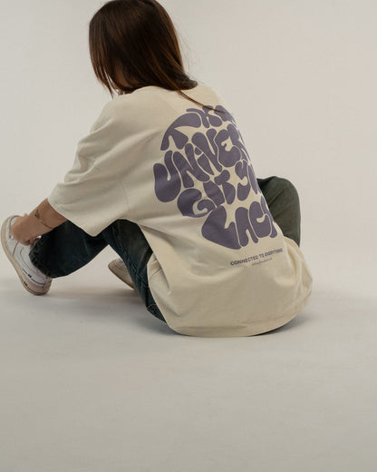 Person sitzt mit überkreuzten Beinen auf dem Boden und trägt ein beigefarbenes T-Shirt mit großem Rückendruck in lila Schrift: „THE UNIVERSE GOT YOUR BACK“. Der Druck ist vollständig sichtbar.