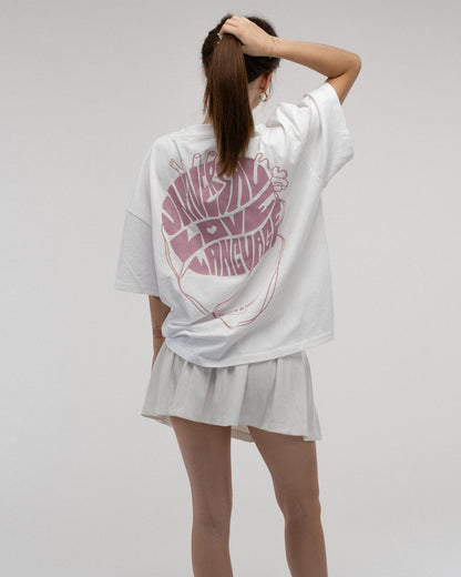 Rückansicht einer stehenden Person mit weißem T-Shirt. Auf dem Rücken ist ein großes, rosafarbenes Motiv in Herzform mit den Worten „UNIVERSAL LOVE LANGUAGE“. Der untere Text lautet „HEARTS IN HARMONY – clothing from planet earth“.