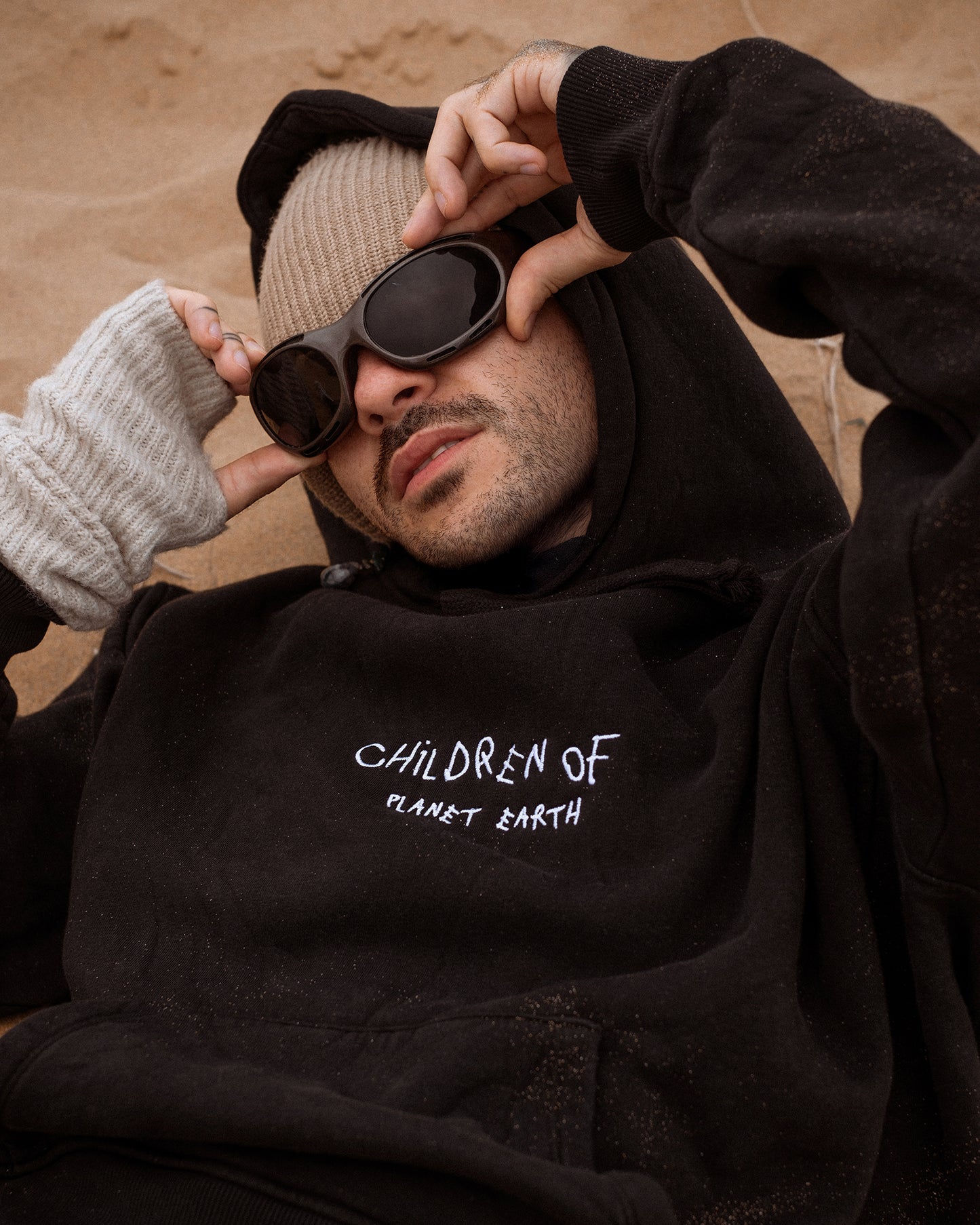 Person liegt entspannt im Sand und trägt einen schwarzen Hoodie mit der weißen Aufschrift „Children of Planet Earth“. Der Look wird durch eine beigefarbene Strickmütze, dunkle Sonnenbrille und fingerlose Strickhandschuhe ergänzt – lässiger Streetwear-Stil mit natürlichem, urbanem Vibe.