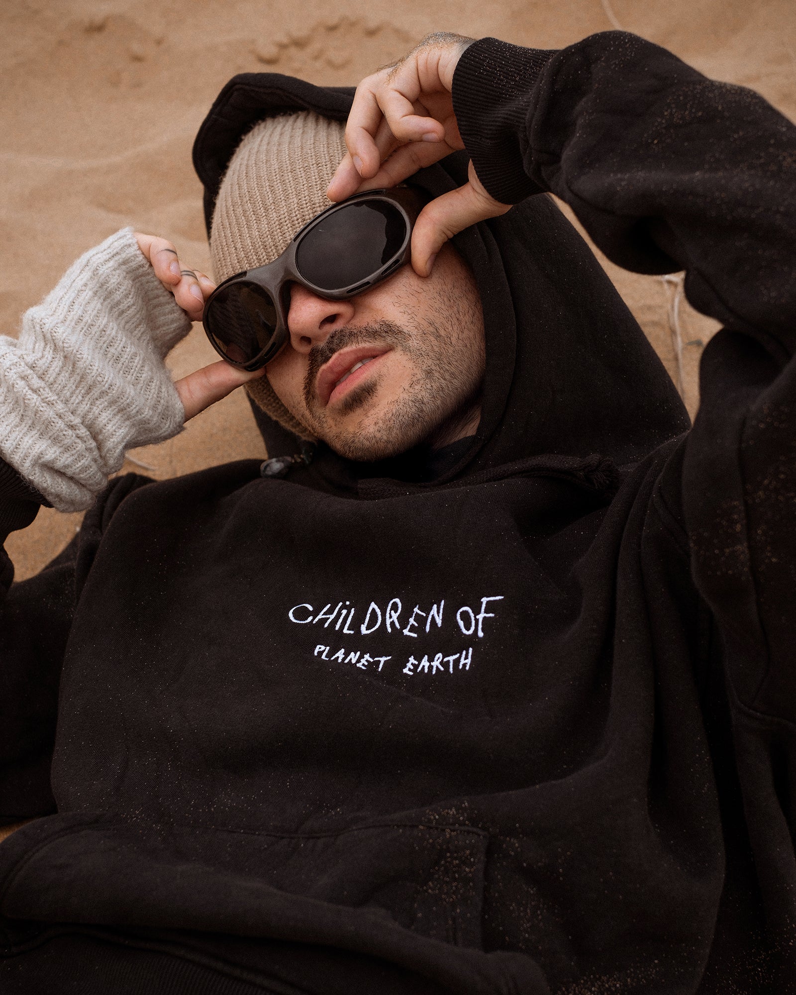 Person liegt entspannt im Sand und trägt einen schwarzen Hoodie mit der weißen Aufschrift „Children of Planet Earth“. Der Look wird durch eine beigefarbene Strickmütze, dunkle Sonnenbrille und fingerlose Strickhandschuhe ergänzt – lässiger Streetwear-Stil mit natürlichem, urbanem Vibe.