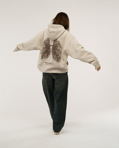 Beiger Kapuzenpullover mit großem Rückenprint in dunkelbraun: eine organische Illustration in Form von Lungenflügeln aus Baumwurzeln. Darunter der Slogan „GOOD SHIT TAKES TIME“. Getragen von einem Model mit ausgebreiteten Armen.