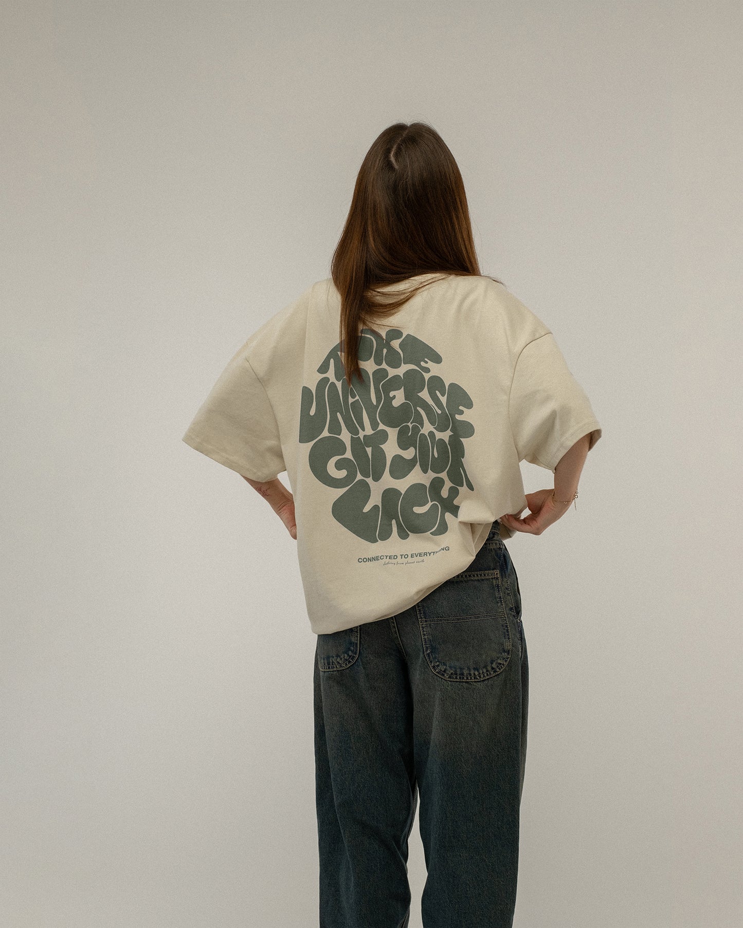 Beiges T-Shirt mit grünem Rückendruck „THE UNIVERSE GOT YOUR BACK“ wird von einer stehenden Person getragen, die sich leicht zur Seite dreht. Der Druck ist vollständig sichtbar.