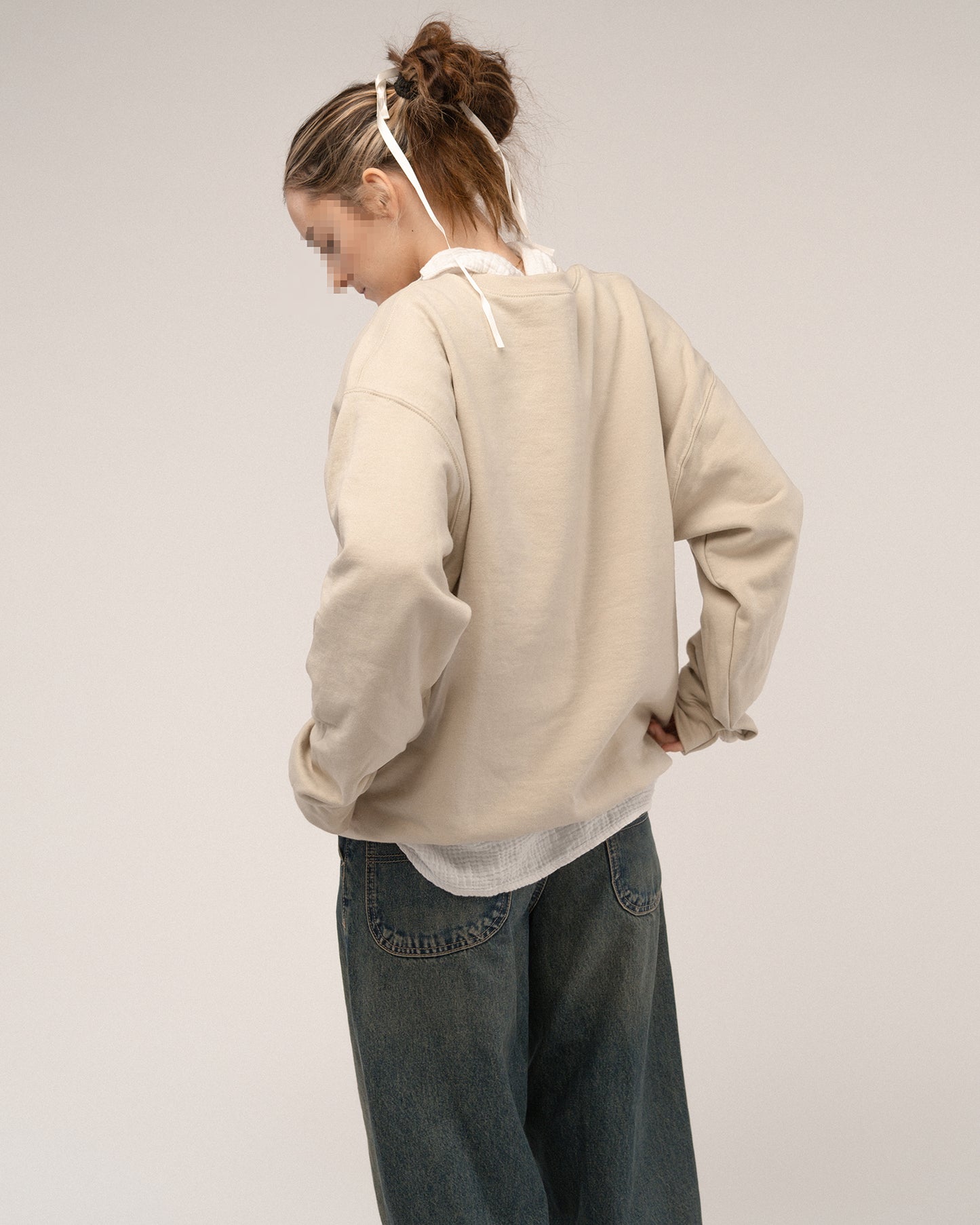 Rückansicht eines beigefarbenen Sweatshirts ohne sichtbares Motiv, getragen über einer weißen Bluse mit lockerer Jeans.