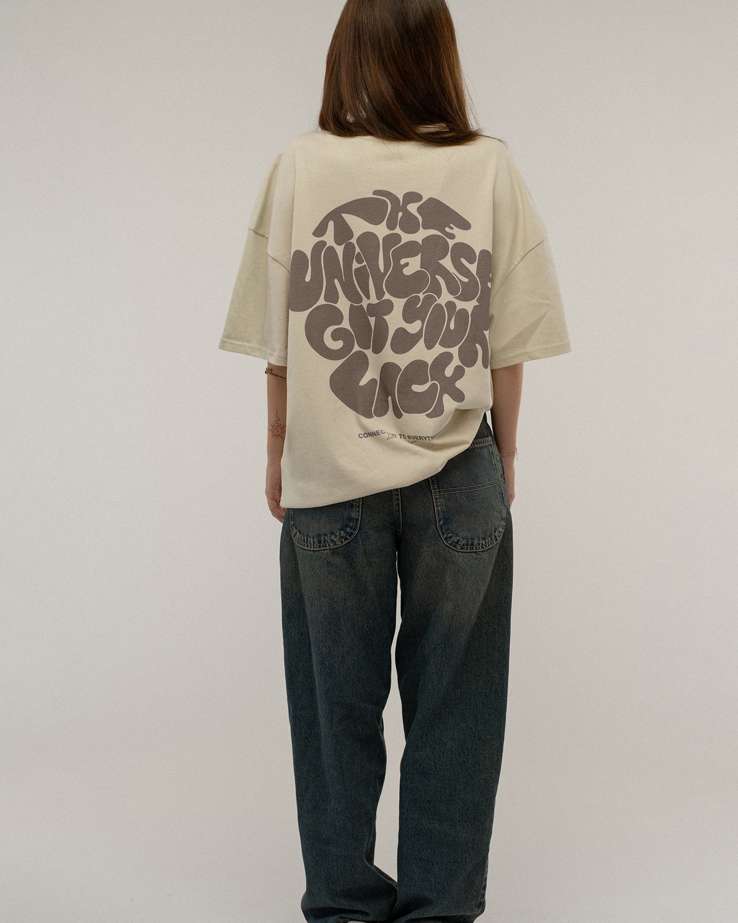 Beiges T-Shirt mit großem Rückendruck in Braun wird von hinten getragen. Die Aufschrift „THE UNIVERSE GOT YOUR BACK“ ist vollständig sichtbar, darunter der Slogan „CONNECTED TO EVERYTHING – clothing from planet earth“.