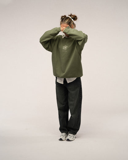 Grünes Sweatshirt mit beiger Stickerei „walking on sunshine“ und Sonnenmotiv auf der Brust, kombiniert mit dunkler Jeans und heller Bluse.