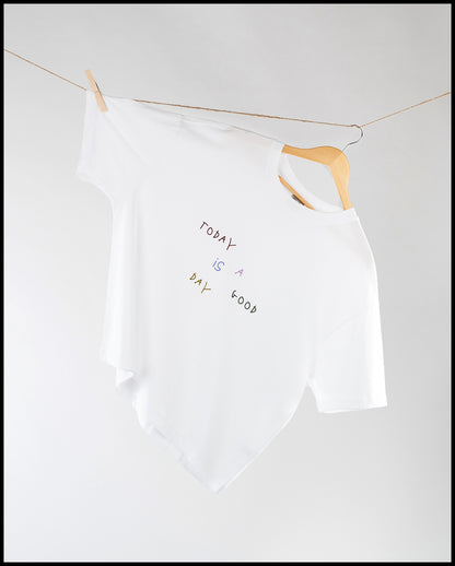  Weißes T-Shirt mit lockerem Schnitt hängt schräg auf einem Holzkleiderbügel an einer Wäscheleine; auf der Vorderseite befindet sich ein bunter, handschriftlicher Schriftzug „Today is a good day“.
