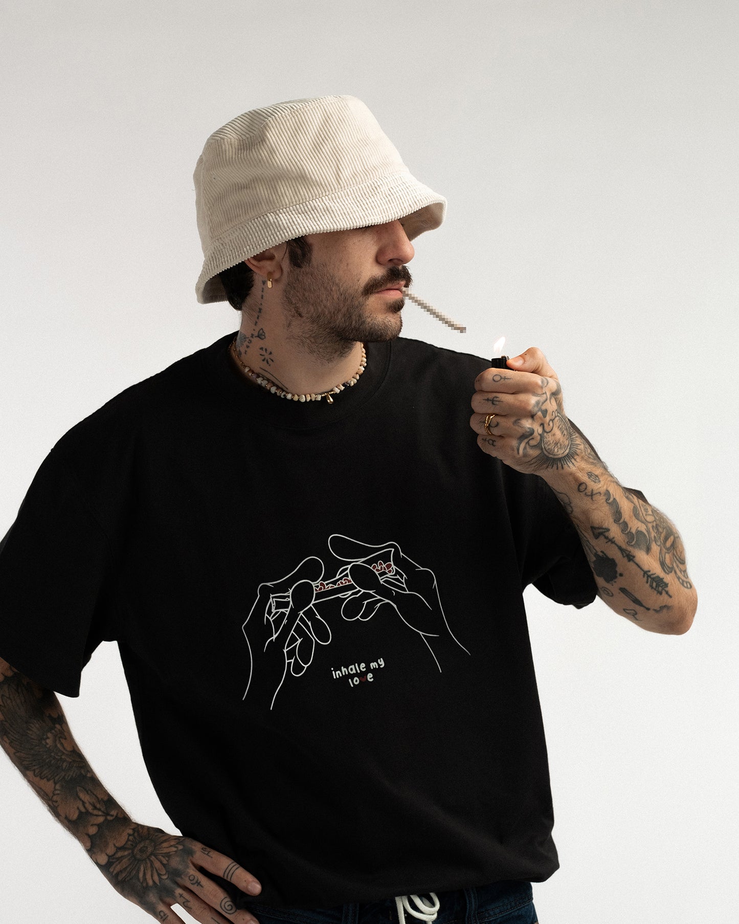 Schwarzes T-Shirt mit weißer Grafik zweier Hände, die einen Joint halten. Die Aufschrift „inhale my love“ ist in geschwungener Schreibschrift unter dem Motiv zu sehen.
