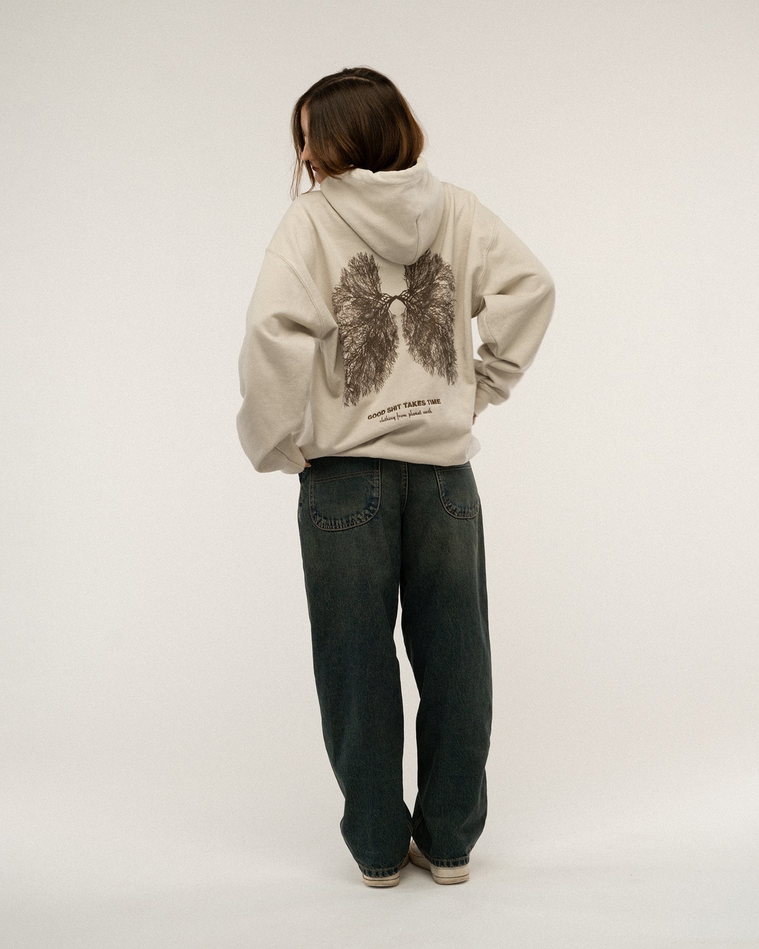Beiger Kapuzenpullover mit großem dunkelbraunem Print auf dem Rücken. Die Illustration zeigt Lungenflügel aus feinen Baumwurzeln mit dem Text „GOOD SHIT TAKES TIME“ darunter. Das Model steht ruhig mit Blick zur Seite.