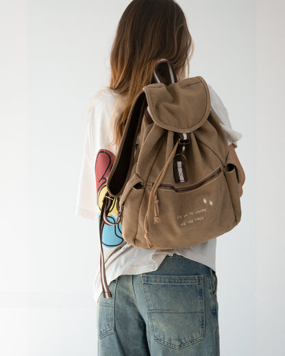 Sandfarbener Canvas-Rucksack am Rücken getragen; gepolsterte Schulterriemen mit braun-weißem Webband, Überschlag mit Haken, Front-Reißverschlussfach mit Stickmotiv.