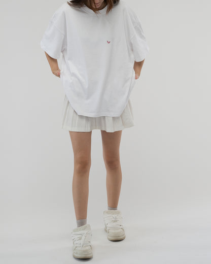 Vorderansicht einer stehenden Person in einem weißen, locker sitzenden T-Shirt. Auf der Brust ist ein kleines, rosafarbenes Herzsymbol abgebildet. Der Rückenaufdruck ist nicht sichtbar.