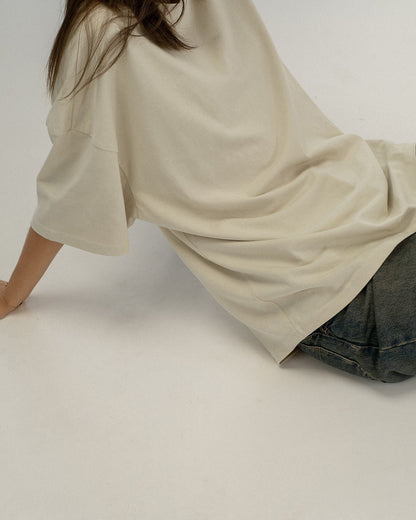 Beiges T-Shirt ohne sichtbaren Druck liegt locker fallend auf dem Rücken einer seitlich ausgestreckten Person. Der Fokus liegt auf dem Material und Schnitt des Shirts.