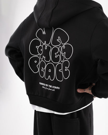 Rückansicht eines schwarzen Hoodies mit auffälligem Rückendruck in weißer Schrift. Großflächig steht ‚WE COME IN PEACE‘ in verspielter, organischer Typografie. Darunter kleinere Texte: ‚GUIDED BY THE STARS‘ und ‚clothing from planet earth‘