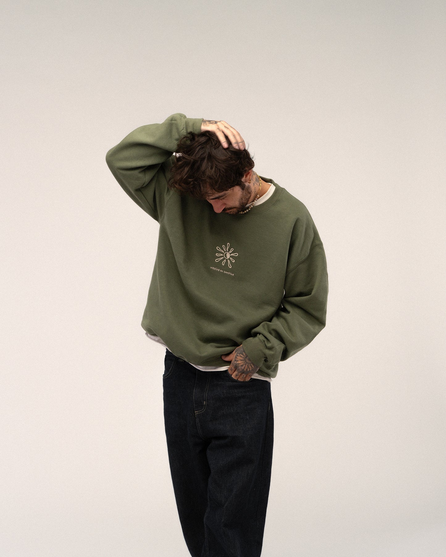 Grünes Sweatshirt mit Stickerei „walking on sunshine“ und Sonne, getragen von einer tätowierten Person in dunkler Jeans.