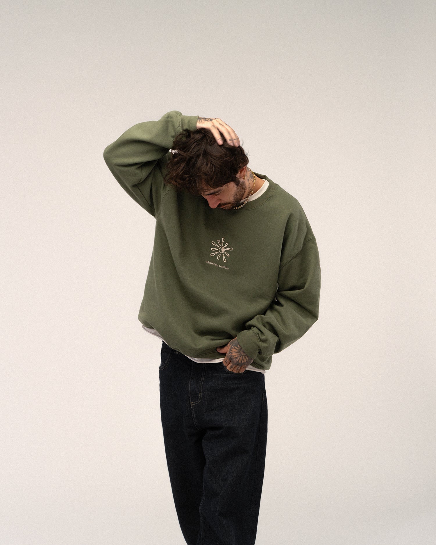 Grünes Sweatshirt mit Stickerei „walking on sunshine“ und Sonne, getragen von einer tätowierten Person in dunkler Jeans.