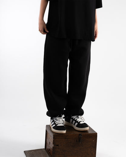 Person steht auf einer kleinen Holzkiste, trägt eine weite schwarze Jogginghose mit elastischem Bund und weiß-schwarzen Sneakern. Vom Oberkörper ist nur ein schwarzes Shirt sichtbar.