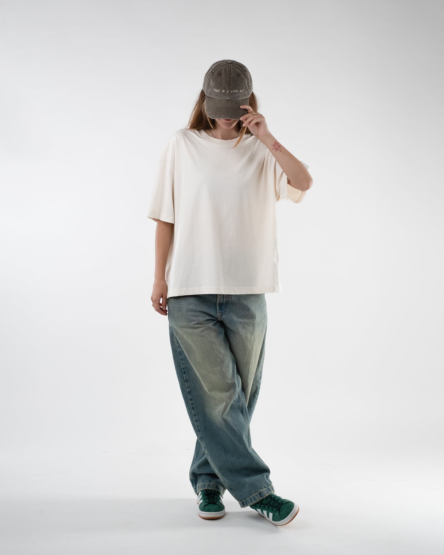 Person mit beigefarbenem T-Shirt in weiter Passform, locker getragen mit Baseballcap, weiter Jeans und grünen Sneakern. Der Blick ist nach unten gerichtet, die Vorderseite des Shirts ist schlicht ohne sichtbaren Aufdruck.