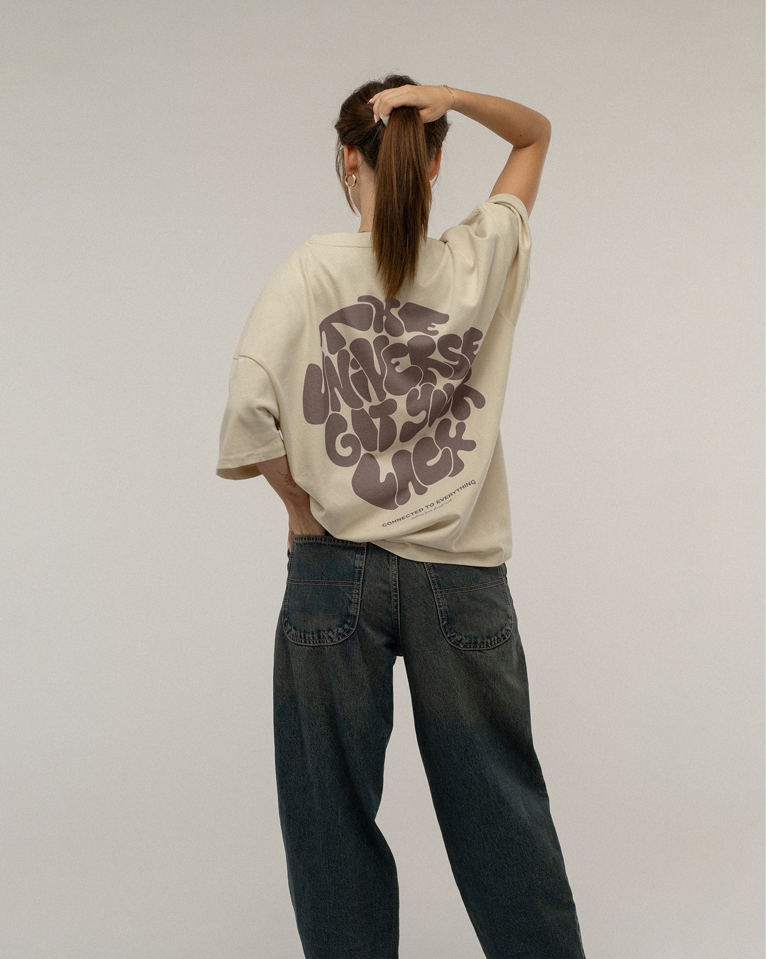 Beiges T-Shirt mit dunkelbraunem Rückendruck wird von hinten getragen. Die Trägerin greift sich in die Haare. Der gesamte Motivtext „THE UNIVERSE GOT YOUR BACK“ mit darunter stehendem Slogan ist sichtbar.
