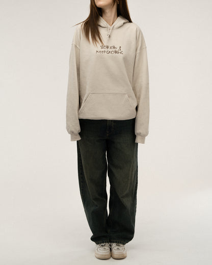 Beiger Kapuzenpullover in Nahaufnahme, Fokus auf die dunkelbraune Bruststickerei „SLOW DOWN & KEEP GROWING“. Sichtbar sind auch Kapuze, Kordeln und der obere Teil der Kängurutasche.