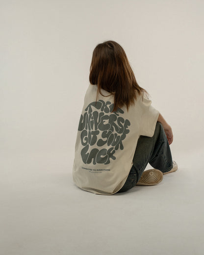 Beiges T-Shirt mit grünem Rückendruck wird von einer sitzenden Person von hinten getragen. Der Schriftzug „THE UNIVERSE GOT YOUR BACK“ ist gut lesbar.