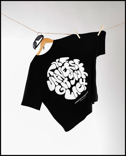 Schwarzes T-Shirt mit weißem Rückseitendruck „The Universe Got Your Back“, hängend an einem Holzkleiderbügel mit Sonnenbrille