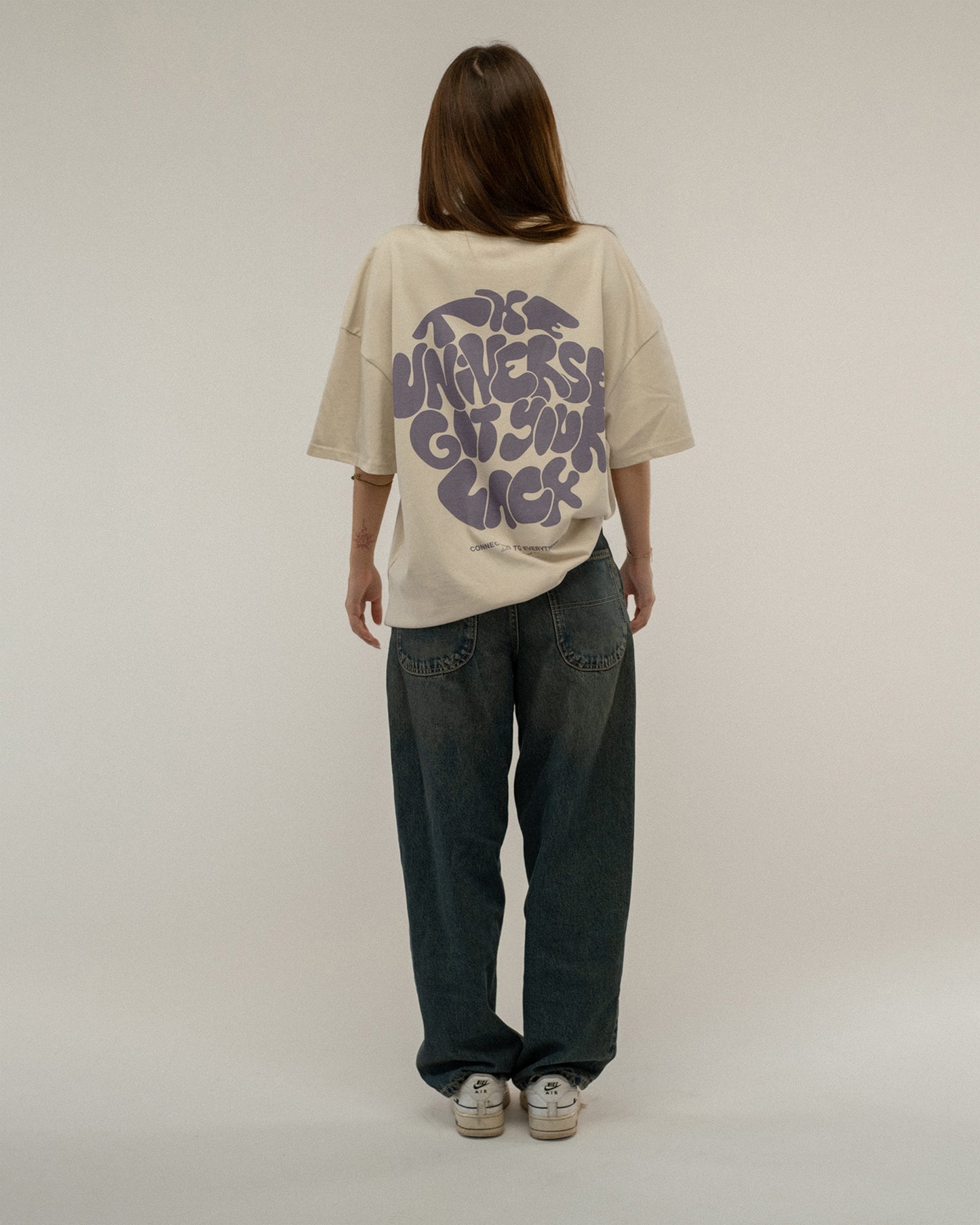 Rückansicht einer stehenden Person mit langen Haaren, die ein beigefarbenes T-Shirt trägt. Der auffällige Rückendruck mit dem Schriftzug „THE UNIVERSE GOT YOUR BACK“ in lila ist vollständig sichtbar.