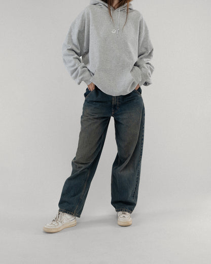 Vorderansicht des grauen Kapuzenpullovers an einer stehenden Person. Auf der Brust ist ein kleines weißes Icon in Form eines stilisierten Blattes abgebildet. Die Hände sind in die Kängurutasche gesteckt, die Kapuze liegt locker am Rücken.