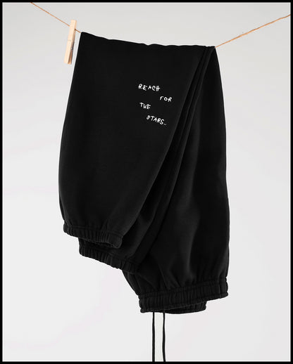 Schwarze Jogginghose hängt locker über einer Wäscheleine mit Holzklammern. Sichtbar ist ein kleiner weißer Sticktext auf dem Oberschenkel: ‚REACH FOR THE STARS..‘