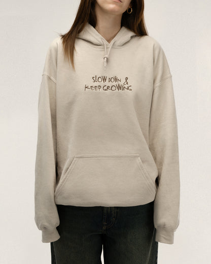 Beiger Kapuzenpullover in lockerer Passform, auf der Vorderseite mit kleiner dunkelbrauner Stickerei über der Brust: „SLOW DOWN & KEEP GROWING“. Das Model steht frontal mit den Händen in der Kängurutasche.