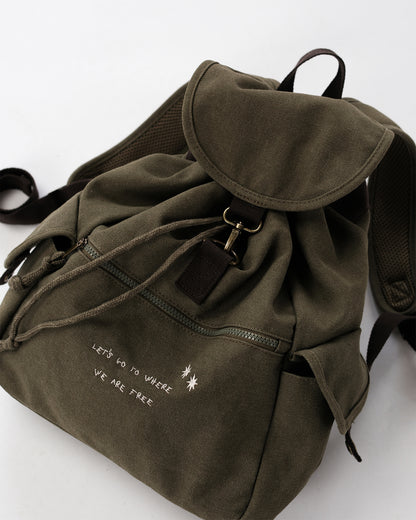 Detailaufnahme olivgrüner Canvas-Rucksack; Überschlag mit Haken und Lederpatch, Kordelzug, Front-Reißverschluss; weiße Stickerei „LET’S GO TO WHERE WE ARE FREE“ mit zwei Sternchen.