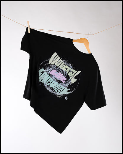 Schwarzes T-Shirt mit farbigem Frontprint, hängend an einem Kleiderbügel auf einer Wäscheleine befestigt. Der Druck zeigt pastellfarbene Schrift im Zentrum einer gesprenkelten Kreisform mit dem Schriftzug ‚UNIVERSAL LOVE LANGUAGE‘ in verspielter Typografie.