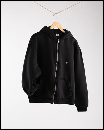 Schwarzer Zip-Hoodie aus Sweatstoff, hängend an einem Holzbügel mit Aufhängung an einer Kordel. Die Jacke hat eine Kapuze, zwei Fronttaschen und einen durchgehenden Reißverschluss. Auf der linken Brust ist ein kleines weißes Peace-Handzeichen aufgestickt.
