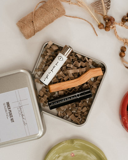 Offene Metallbox mit der Aufschrift „INNER PEACE KIT“, gefüllt mit braunem Verpackungspapier, einem weißen Feuerzeug mit der Aufschrift „a light to guide you“, einem schwarzen Feuerzeug mit der Aufschrift „i’m not scared of the dark“ und einem Palo Santo Holzstück. Daneben Deko mit Bast, Federn und Perlen sowie bunte Keramikteller.