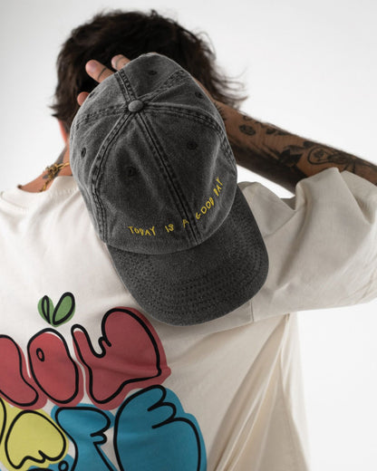 Rückansicht einer schwarzen Cap mit gelber Stickerei „Today is a good day“, getragen auf dem Rücken eines T-Shirts mit buntem Print.
