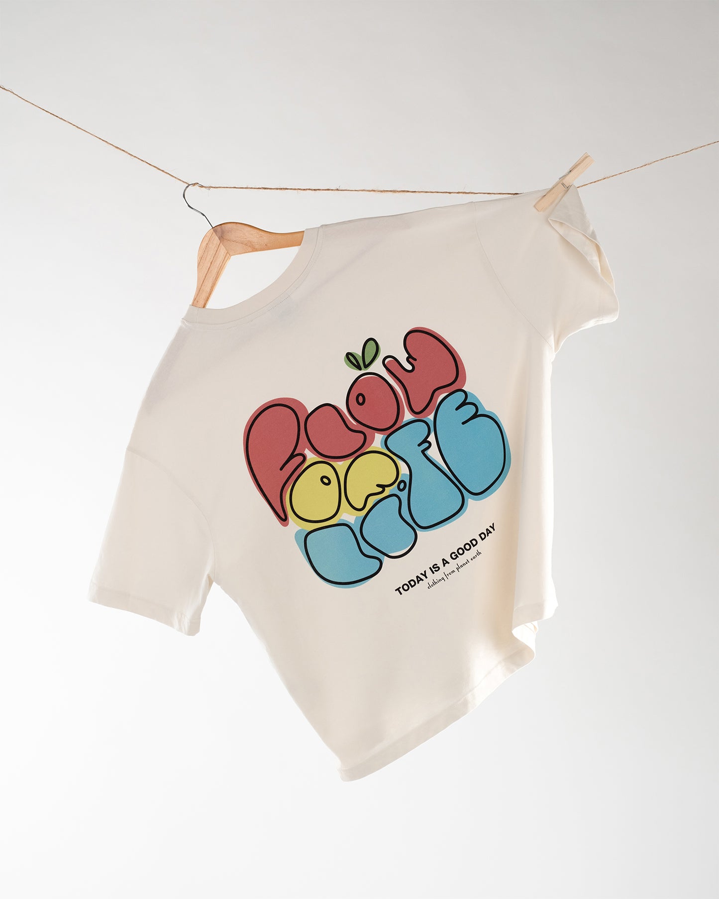 Ein cremefarbenes T-Shirt hängt schräg an einem Kleiderbügel, der an einer Wäscheleine befestigt ist. Auf dem Rücken des Shirts befindet sich ein bunter, verspielter Print mit der Aufschrift „Flow of Life“ in großen Lettern, gestaltet in Rot, Gelb und Blau. Darunter steht in kleiner Schrift: „TODAY IS A GOOD DAY – clothing from planet earth“. Das T-Shirt vermittelt durch Design und Farbe einen positiven, lebensfrohen Eindruck.