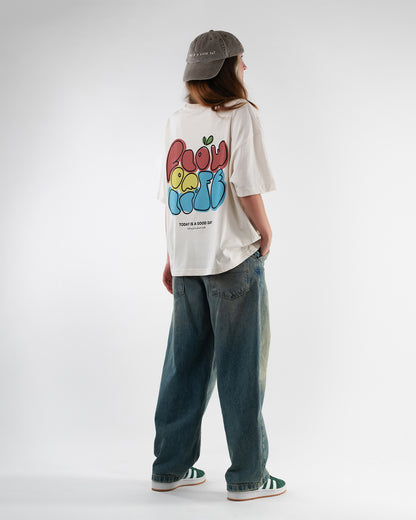Eine Person trägt ein locker sitzendes, cremefarbenes T-Shirt mit großem Rückendruck, der in bunten, runden Buchstaben „Flow of Life“ zeigt. Darunter steht „TODAY IS A GOOD DAY – clothing from planet earth“. Die Person steht mit dem Rücken zur Kamera, trägt eine graue Cap, eine weite Jeans und grüne Sneaker. Das T-Shirt wirkt lässig, freundlich und jugendlich.