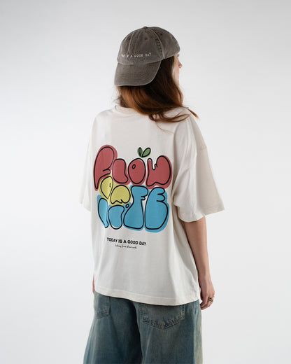 Nahaufnahme einer Person von hinten, die ein cremefarbenes Oversize-T-Shirt mit einem farbenfrohen Print trägt. Der Aufdruck zeigt die Worte „Flow of Life“ in rot, gelb und blau mit kleinen grünen Blättern über dem „L“. Darunter ist der motivierende Spruch „TODAY IS A GOOD DAY – clothing from planet earth“ zu lesen. Der Look unterstreicht einen fröhlichen, urbanen Stil mit nachhaltiger Botschaft.