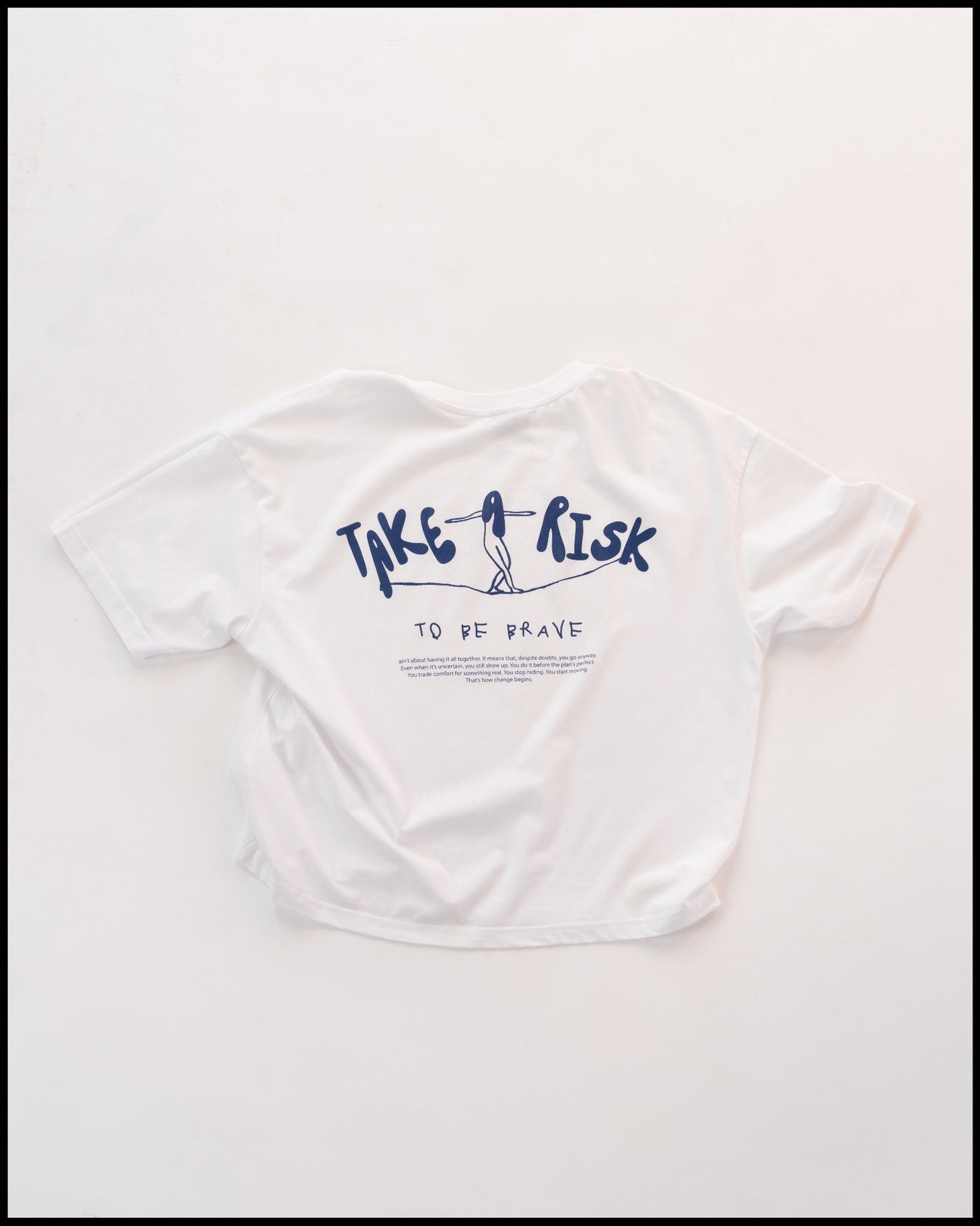 Weißes T-Shirt mit dunkelblauem Aufdruck „TAKE A RISK“ auf der Rückseite, darunter der Schriftzug „TO BE BRAVE“. Die Illustration zeigt eine Figur, die auf einer Linie balanciert. Minimalistisches und inspirierendes Design im modernen Stil.