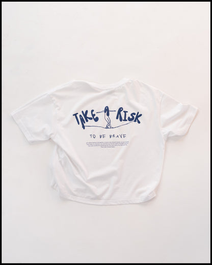 Weißes T-Shirt mit dunkelblauem Aufdruck „TAKE A RISK“ auf der Rückseite, darunter der Schriftzug „TO BE BRAVE“. Die Illustration zeigt eine Figur, die auf einer Linie balanciert. Minimalistisches und inspirierendes Design im modernen Stil.