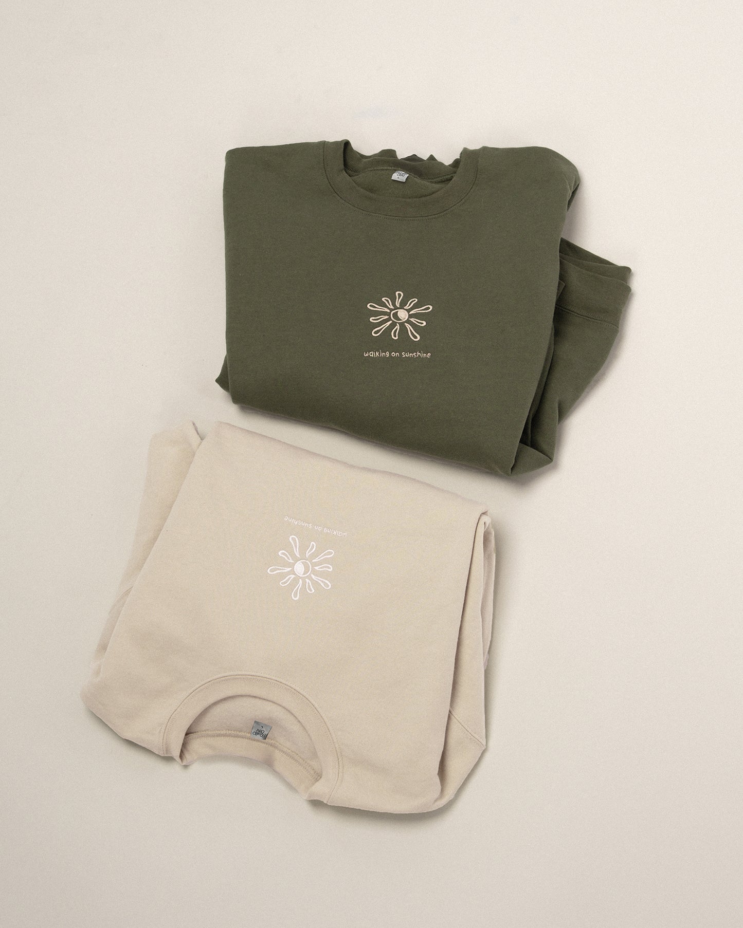Zwei übereinander gelegte Sweatshirts in Beige und Grün, beide mit gesticktem Sonnenmotiv und dem Text „walking on sunshine“.