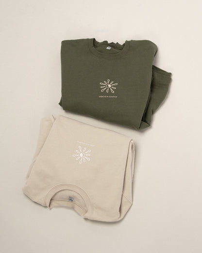 Zwei übereinander gelegte Sweatshirts in Beige und Grün, beide mit gesticktem Sonnenmotiv und dem Text „walking on sunshine“.