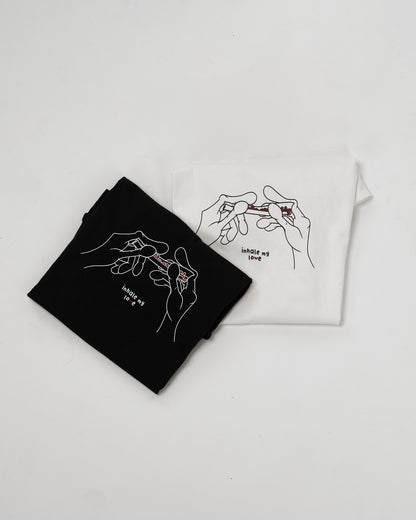 Zwei zusammengefaltete T-Shirts, eins in Schwarz und eins in Weiß, beide mit derselben grafischen Illustration: zwei stilisierte Hände drehen einen Joint, mit der Aufschrift „inhale my love“ darunter.